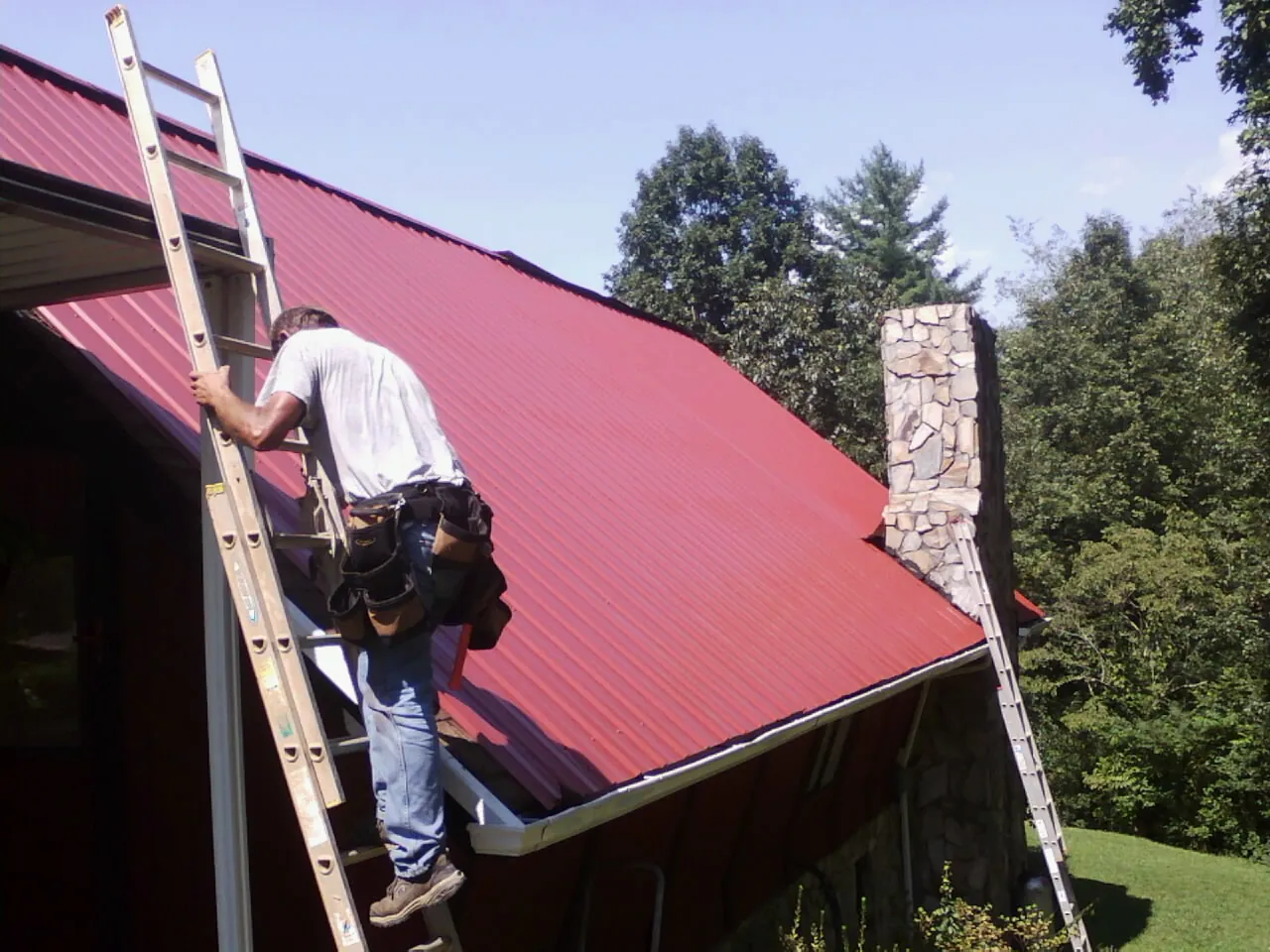 Roofer on a ladder beside a red metal roof and stone chimney — Soffit & Fascia Repair by Roofing Brothers in Enterprise